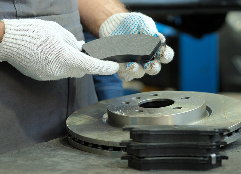 Gloved hands hold a new brake pad over a desk with a brake disc and more brake pads.